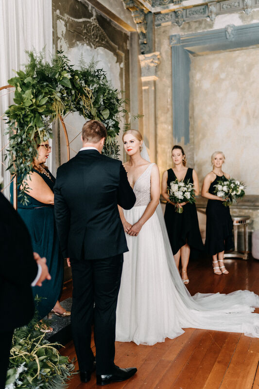 Bride and groom standing under a green foliage arbour at the George Ballroom with bridesmaids in black dresses looking on 