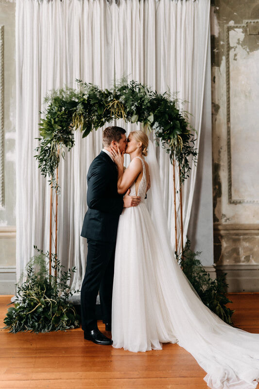 Bride and groom share their first kiss under green foliage arch at the George Ballroom