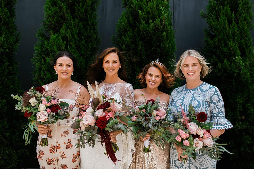 Bride and her bridesmaids holding wild and romantic flower bouquets of pampas grass, eucalyptus, amaranthus and roses in burgundy, silver and blush tones. Bridesmaids are in mismatched dresses of different colours and patterns. 