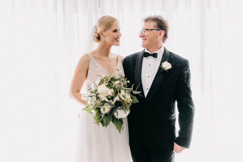 Bride holding wedding bouquet of white peonies and green foliage standing next to her father who wears a black tuxedo and white rose buttonhole 