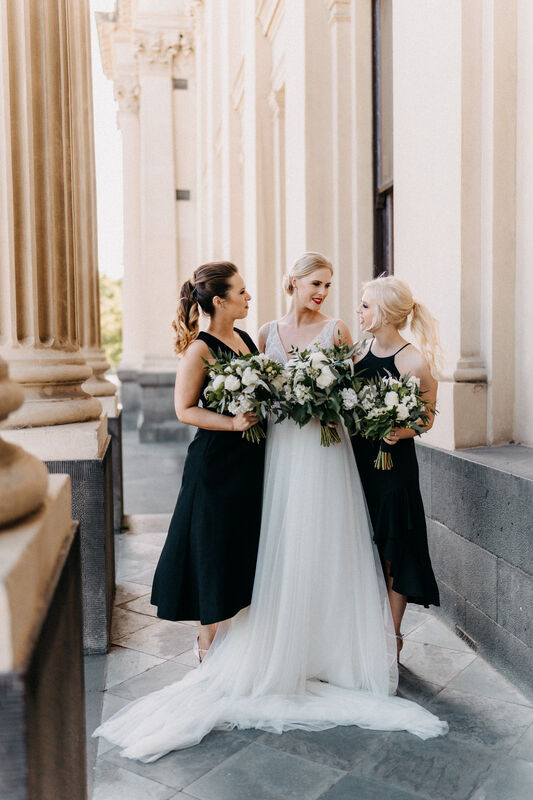Bride and her bridesmaids in black dresses holding flower bouquets of white peonies and green foliage