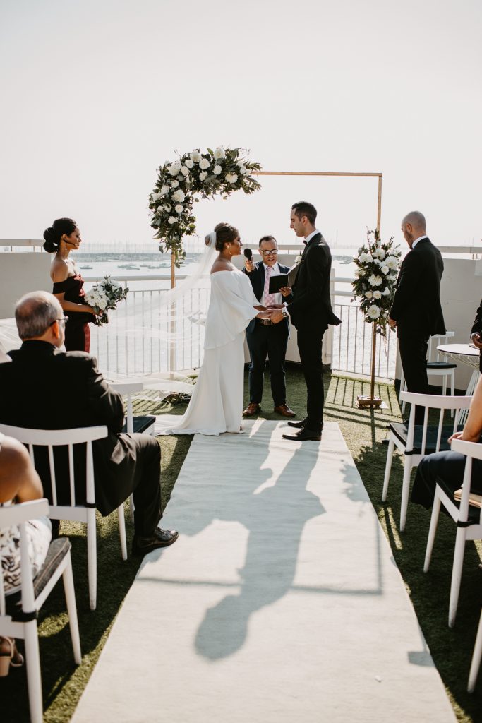 Bride and groom standing under wedding arbour decorated with white and green flowers at Harbour Room St Kilda with views of the beach