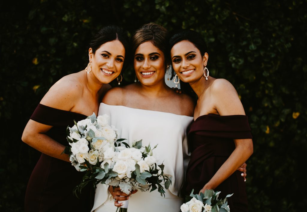 Bride and bridesmaids in black dresses holding flower bouquets of white and green flowers featuring roses and eucalyptus leaves. 