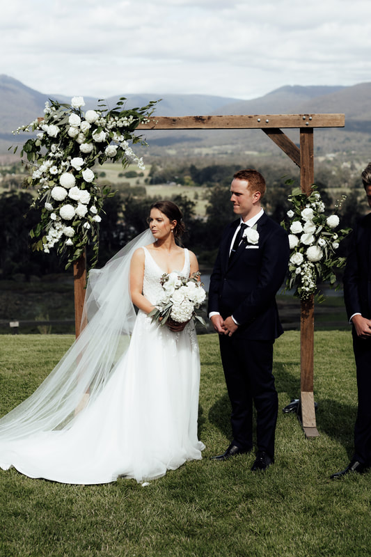 Bride and groom standing under wooden wedding arbour at The Riverstone Estate, Coldstream. The arbour is decorated in the left corner with white and green flowers. 