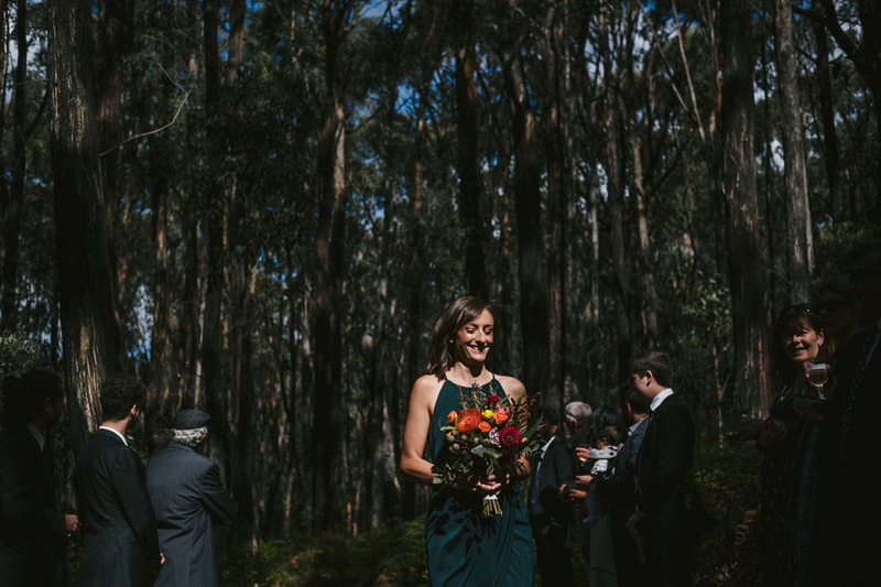 Bridesmaid walks down the aisle in teal dress holding rustic flowers featuring orange and yellow natives. 