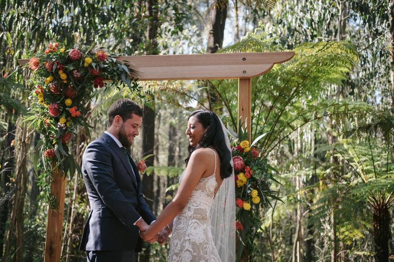 Bride and groom stand under a rustic arbour decorated in the corner with native flowers in bright colours. 