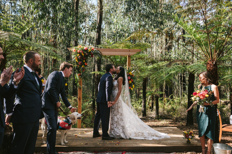 Bride and groom share their first kiss under the arbour at Forest Weddings Kinglake decorated with bright native flowers