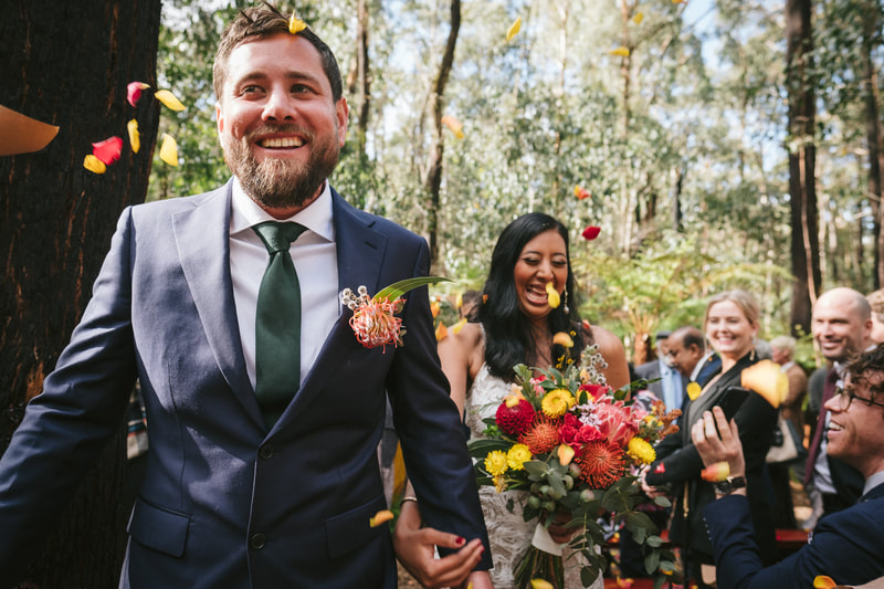 Bride and groom walk back down the aisle after their wedding with colourful rose petals being tossed at them. Bride holds a bright yellow and orange native flower bouquet. 