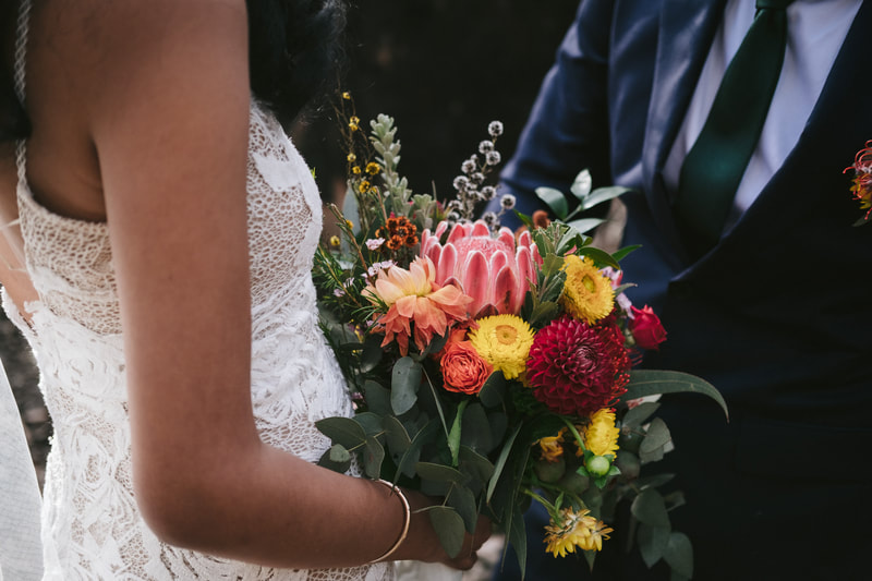 Bright wedding bouquet of proteas, strawflower, eucalyptus and roses in reds, orange and yellow. 