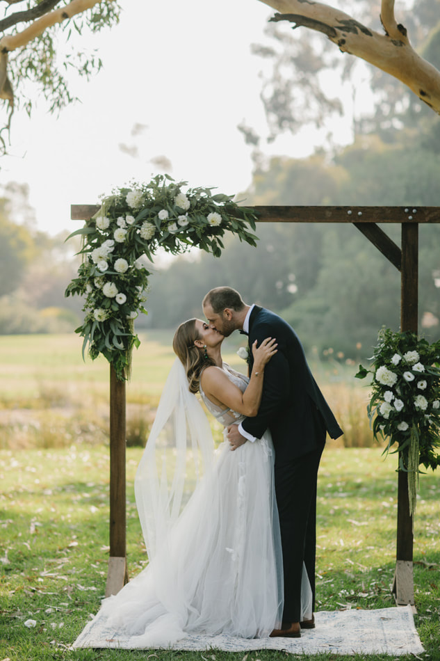 Bride and groom kiss under a wedding arbour made of wood and decorated with rustic green and white flowers including eucalyptus, roses, dahlias. 