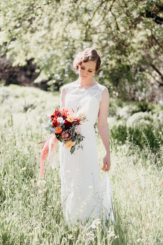 Bride standing in meadow holding a flower bouquet of roses and proteas in burnt orange and burgundy