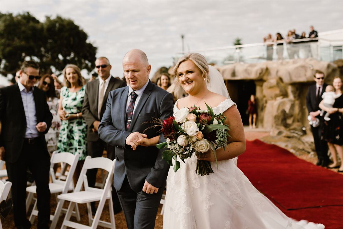 Bride walks down the aisle at Russo Estate with bouquet of burgundy, white and green native flowers