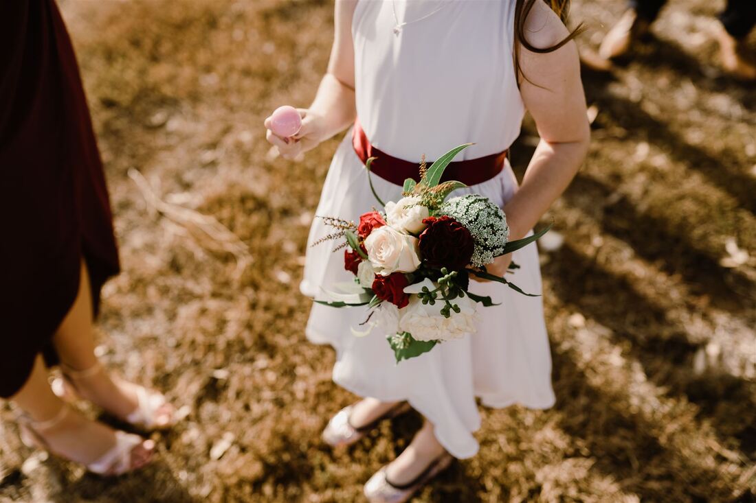Flower girl holding small bouquet of flowers in burgundy, cream and green. 