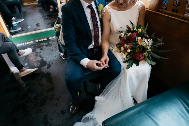 Bride and groom ride a Melbourne tram on their wedding day. Bride is holding a colourful native flower bouquet. 