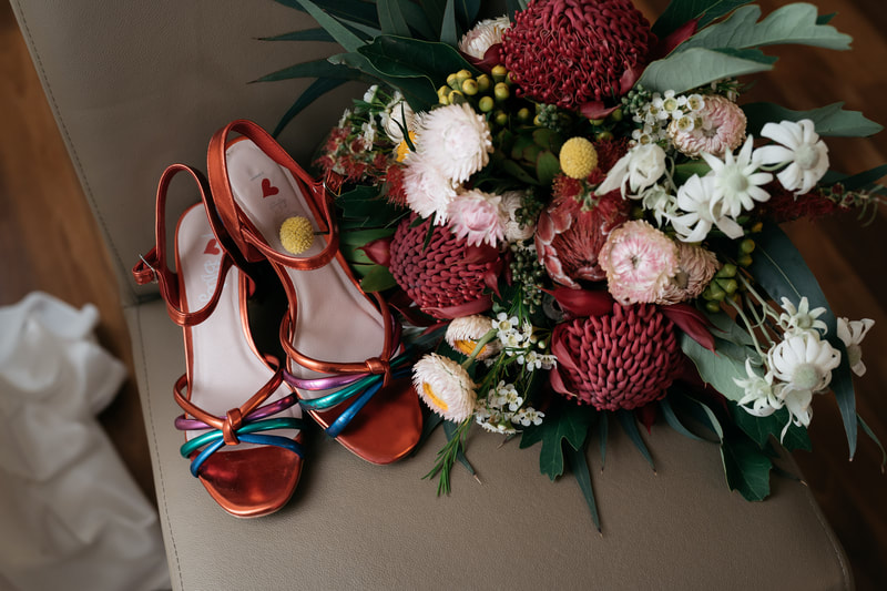 Bridal bouquet of native waratah, proteas, billy buttons, flannel flower and strawflower placed next to bride's colourful wedding shoes. 