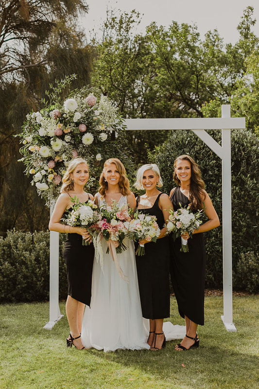 Bride and her bridesmaids stand under a white wood wedding arbour, decorated with flowers in shades of white, green and warm pink. 