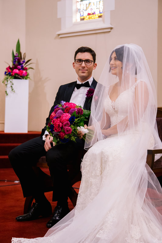 Bride and groom sitting in a church on their wedding day with bright jewel toned flowers. 