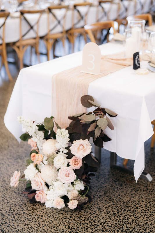 Floral arrangement placed at the base of long reception table, with white and pink flowers and dark foliage. 