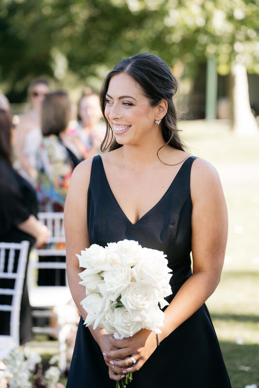 Bridesmaid walks down the aisle in a black dress holding a bouquet of white reflexed roses. 
