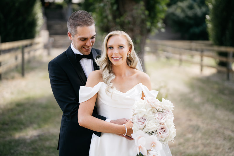 Bride and groom standing in field, bride holds a bouquet of white and blush roses with an orchid. 
